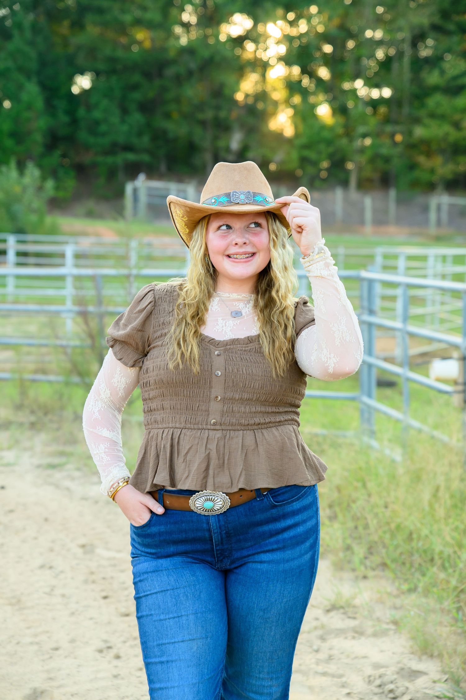 A smiling blonde woman in a cowboy hat, olive top, and jeans poses outdoors near a ranch fence.