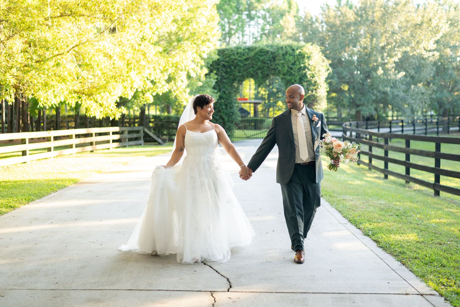 Bride and groom walking hand in hand on a sunlit path surrounded by lush greenery and wooden fences.
