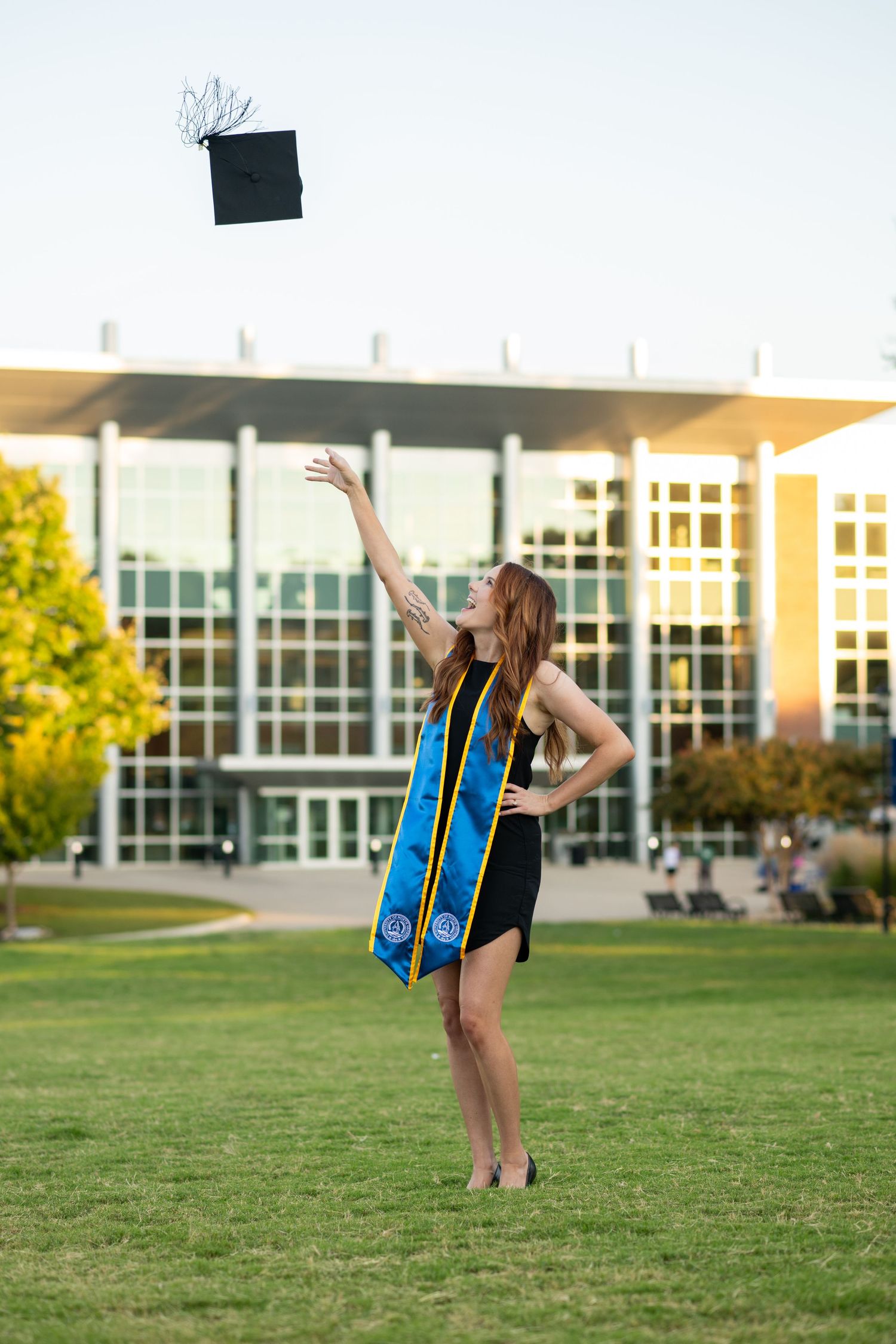 A young woman in graduation attire tosses her cap in the air on a green campus lawn.