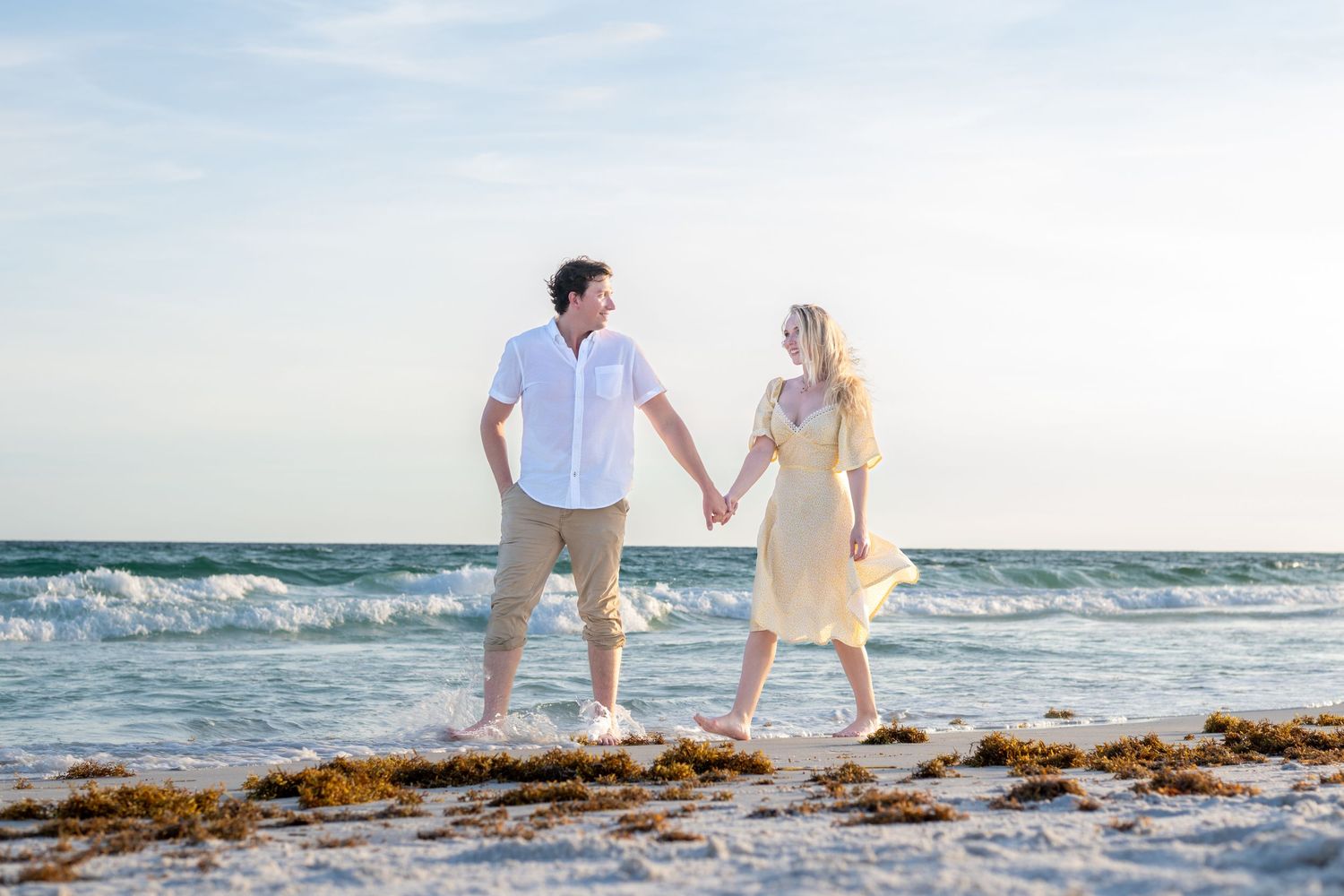 A couple holds hands and gazes at each other while walking barefoot on a sandy beach near ocean waves.