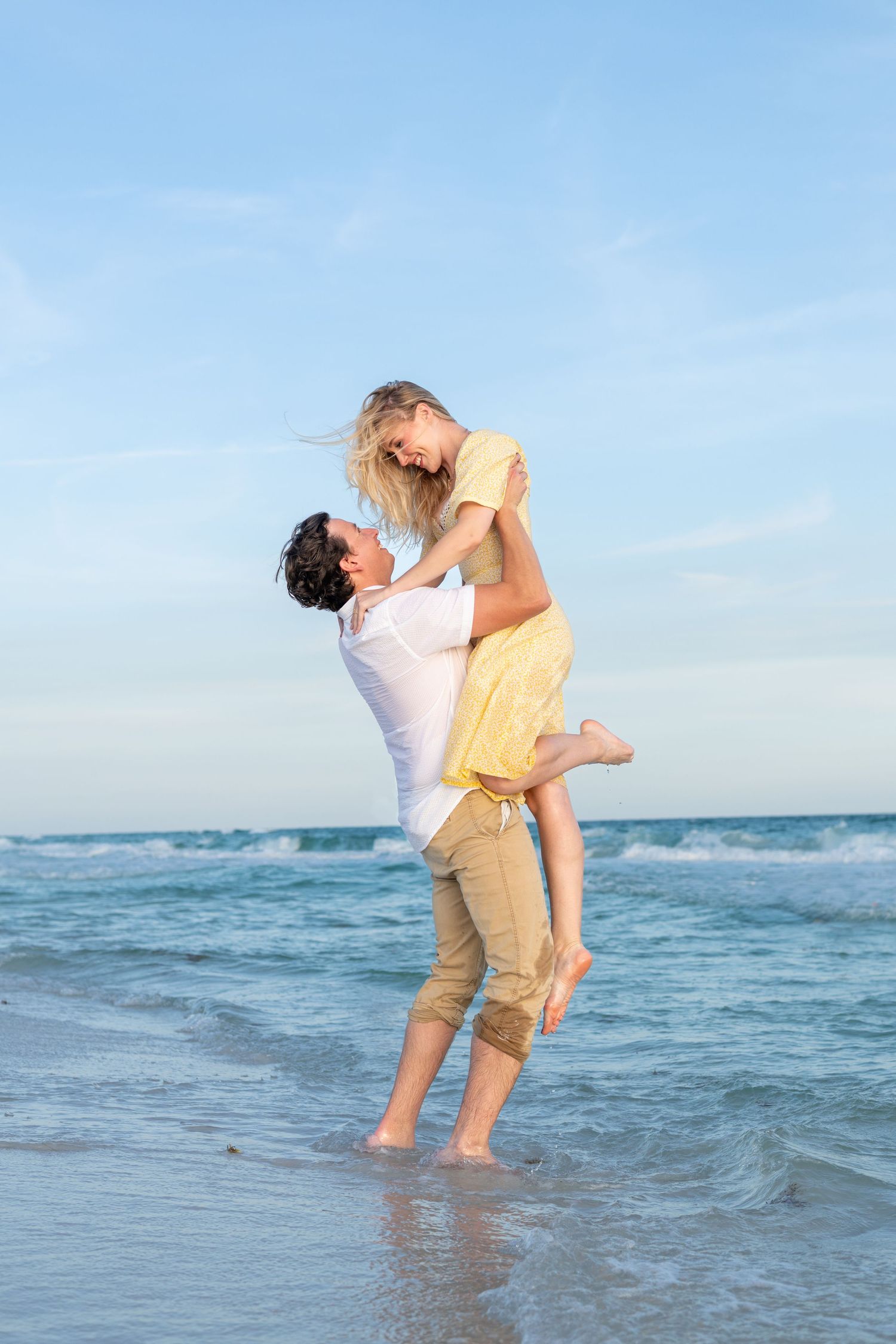 A joyful couple embracing on a beach, man lifting woman in yellow dress, ocean waves in background.