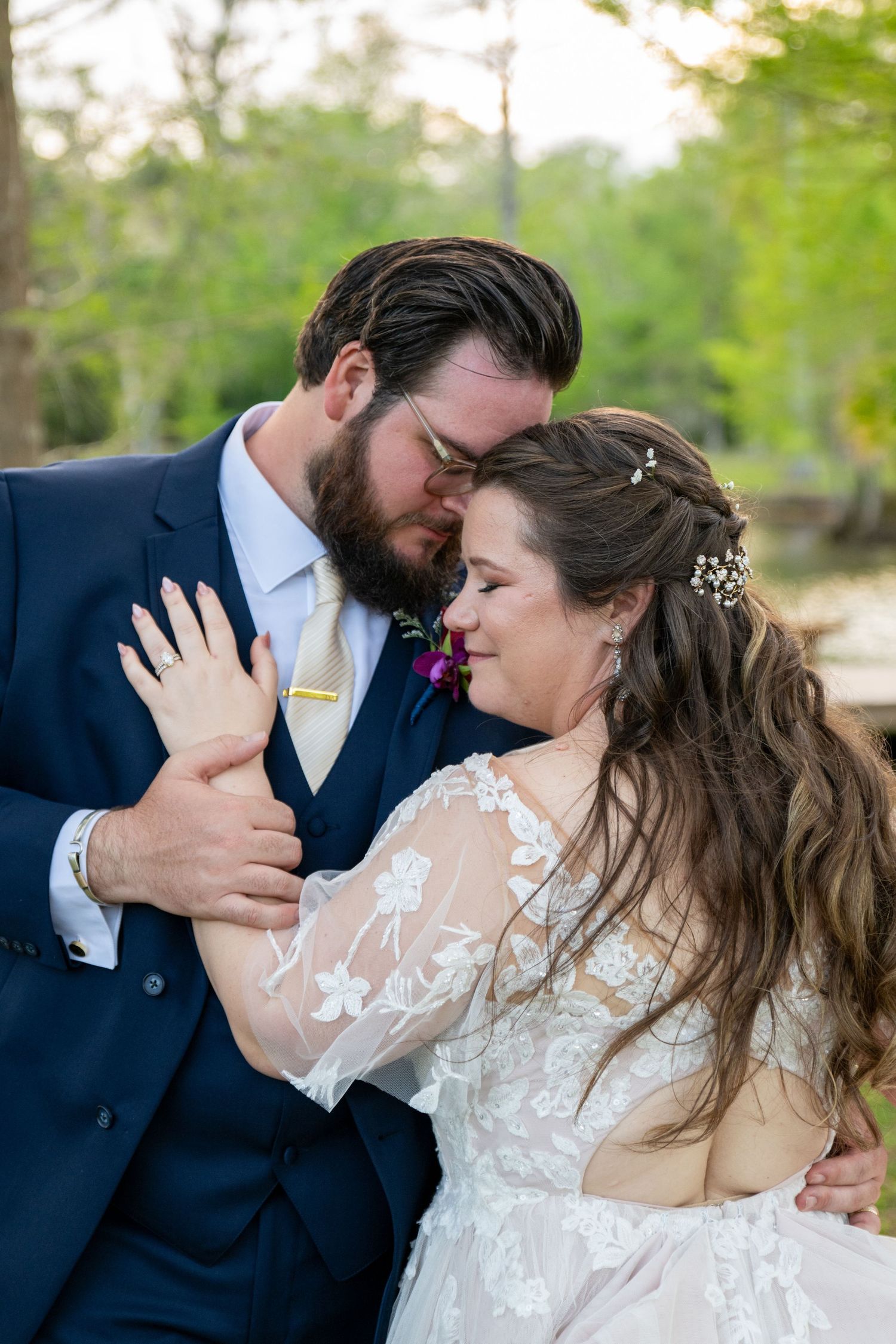 Bearded groom in navy suit tenderly embraces bride in lace gown in a wooded outdoor setting.