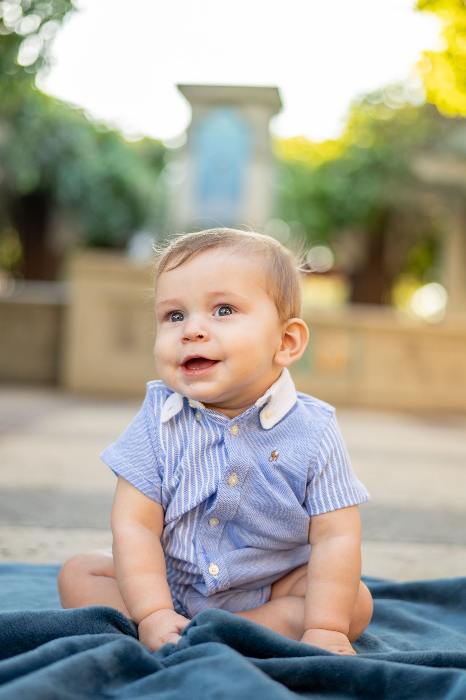 Adorable smiling baby in a blue striped romper sitting on a blanket outdoors.