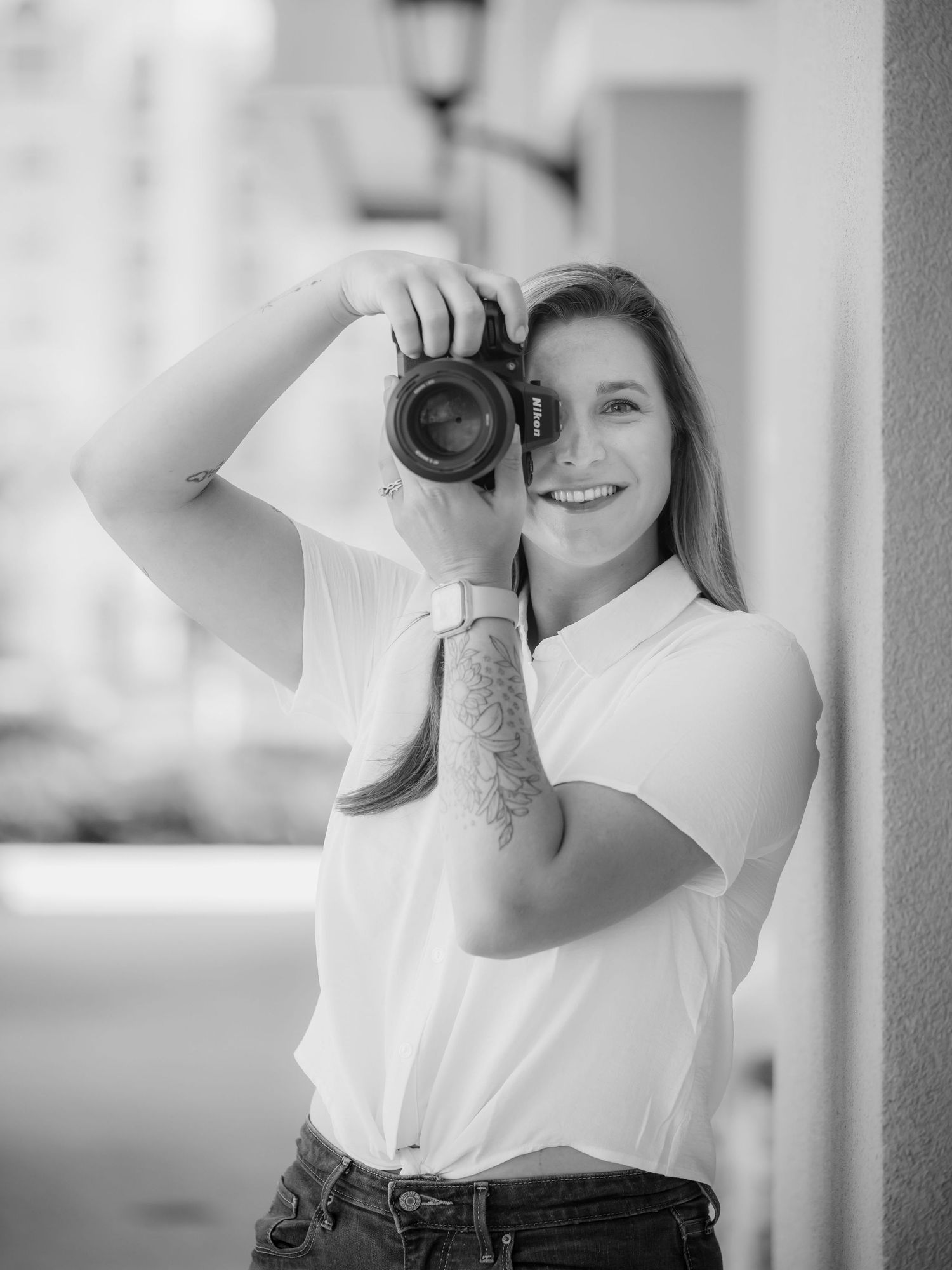 Smiling woman holding a Nikon camera up to her face, wearing a white shirt, with a tattoo on her arm, in black and white.