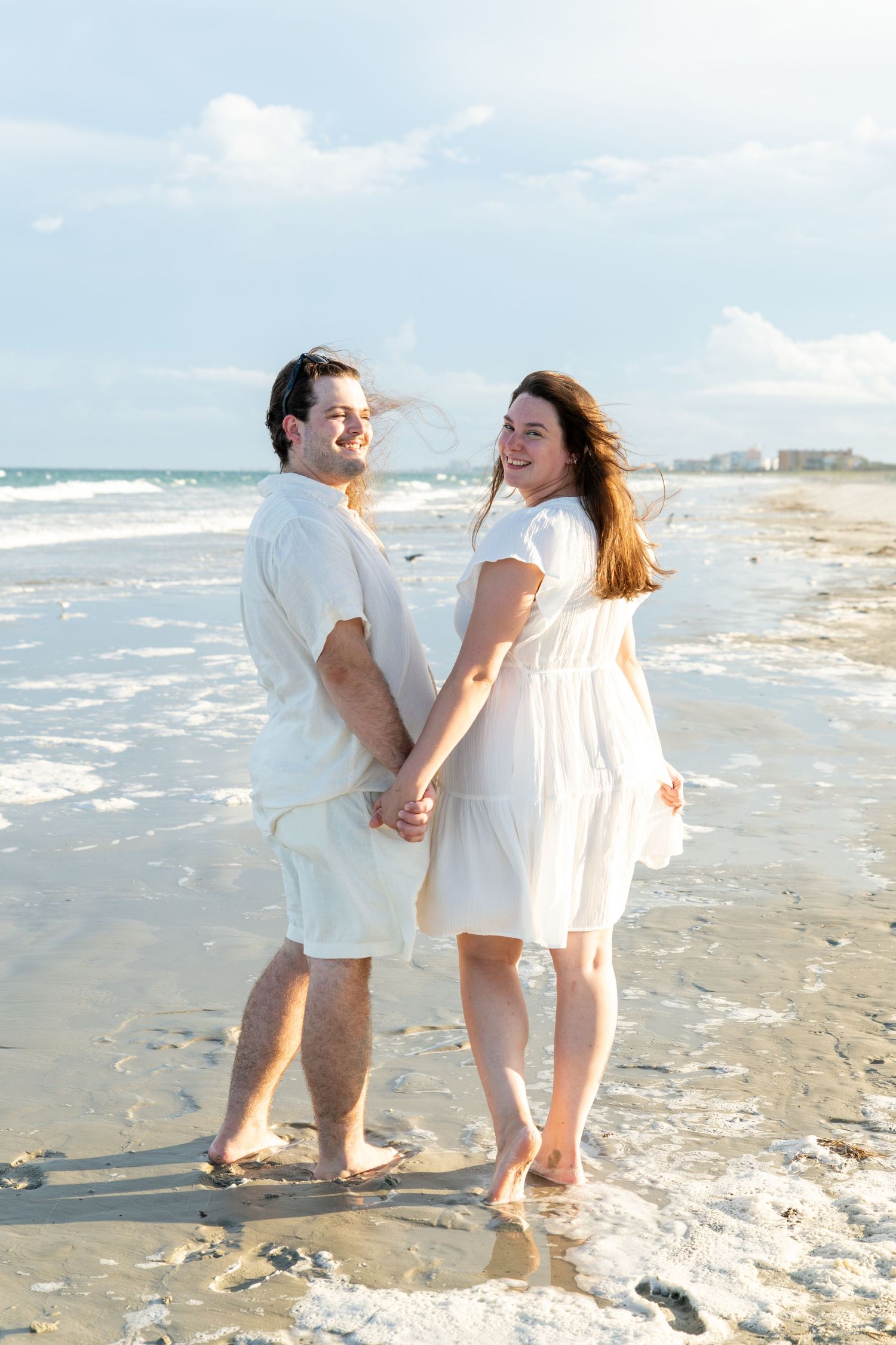 A smiling couple in matching white outfits holds hands on a sunny beach with ocean waves behind them.