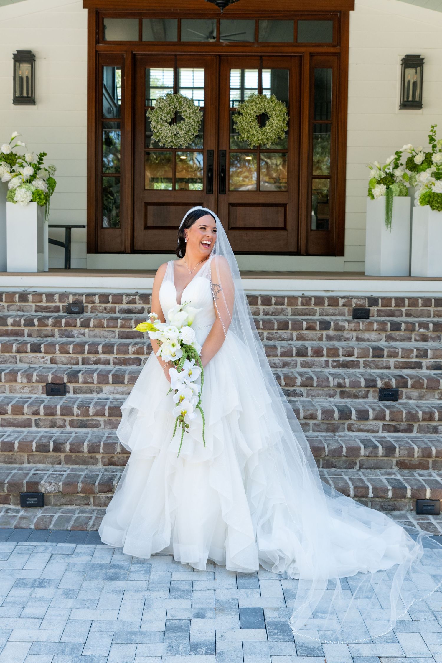 Bride in white layered gown and veil holds white floral bouquet on brick steps before elegant wooden double doors.