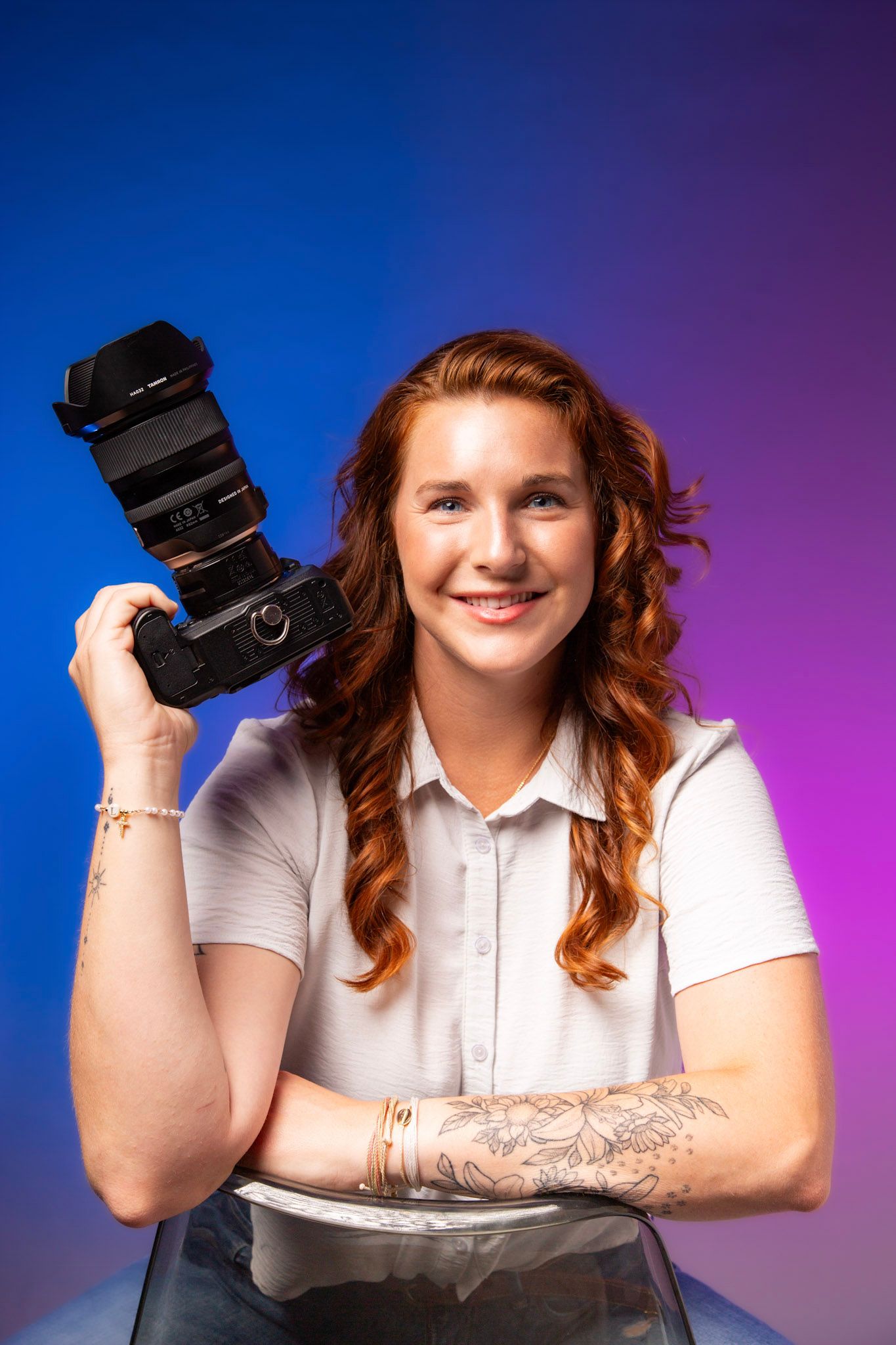 Smiling redhead woman holding a professional DSLR camera with tattoos on her arm against a blue and purple background.