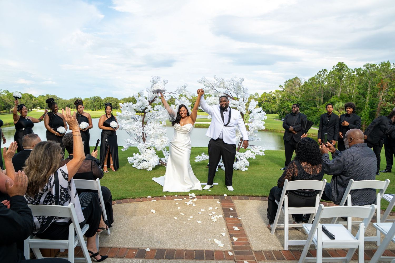 Joyful newlyweds celebrate at outdoor wedding ceremony with white floral arch, guests cheering and rose petals on brick aisle.