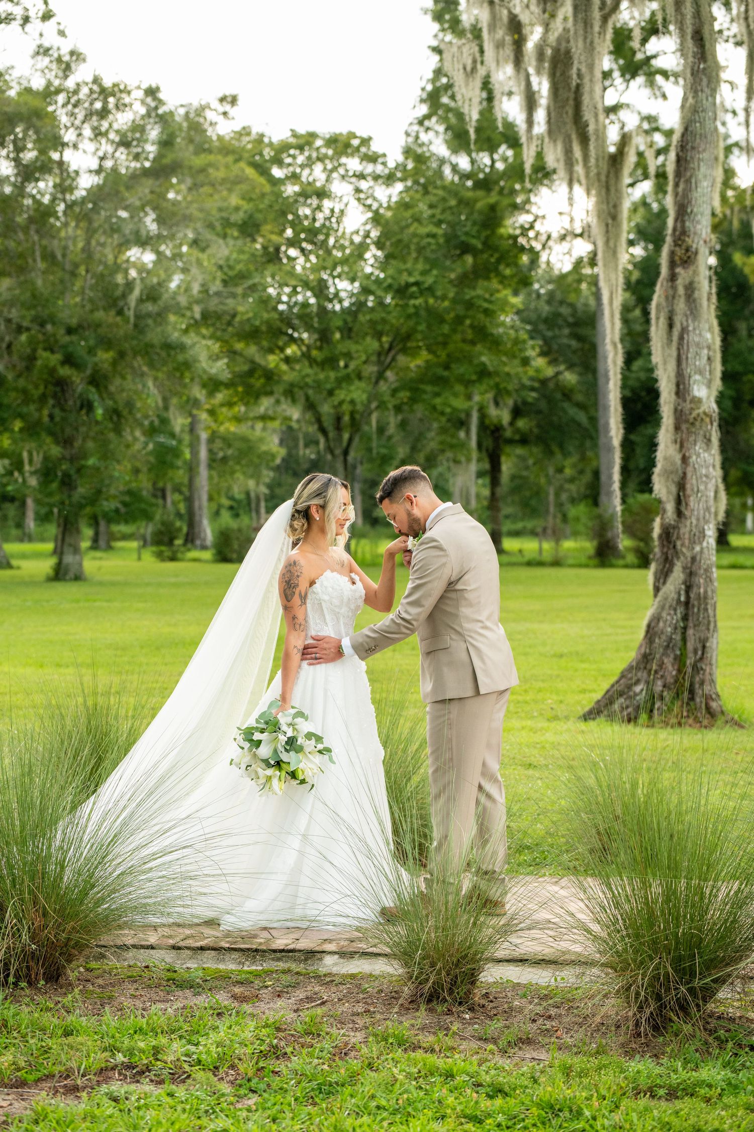 Bride and groom share a romantic moment on a garden path surrounded by lush greenery and Spanish moss-draped trees.