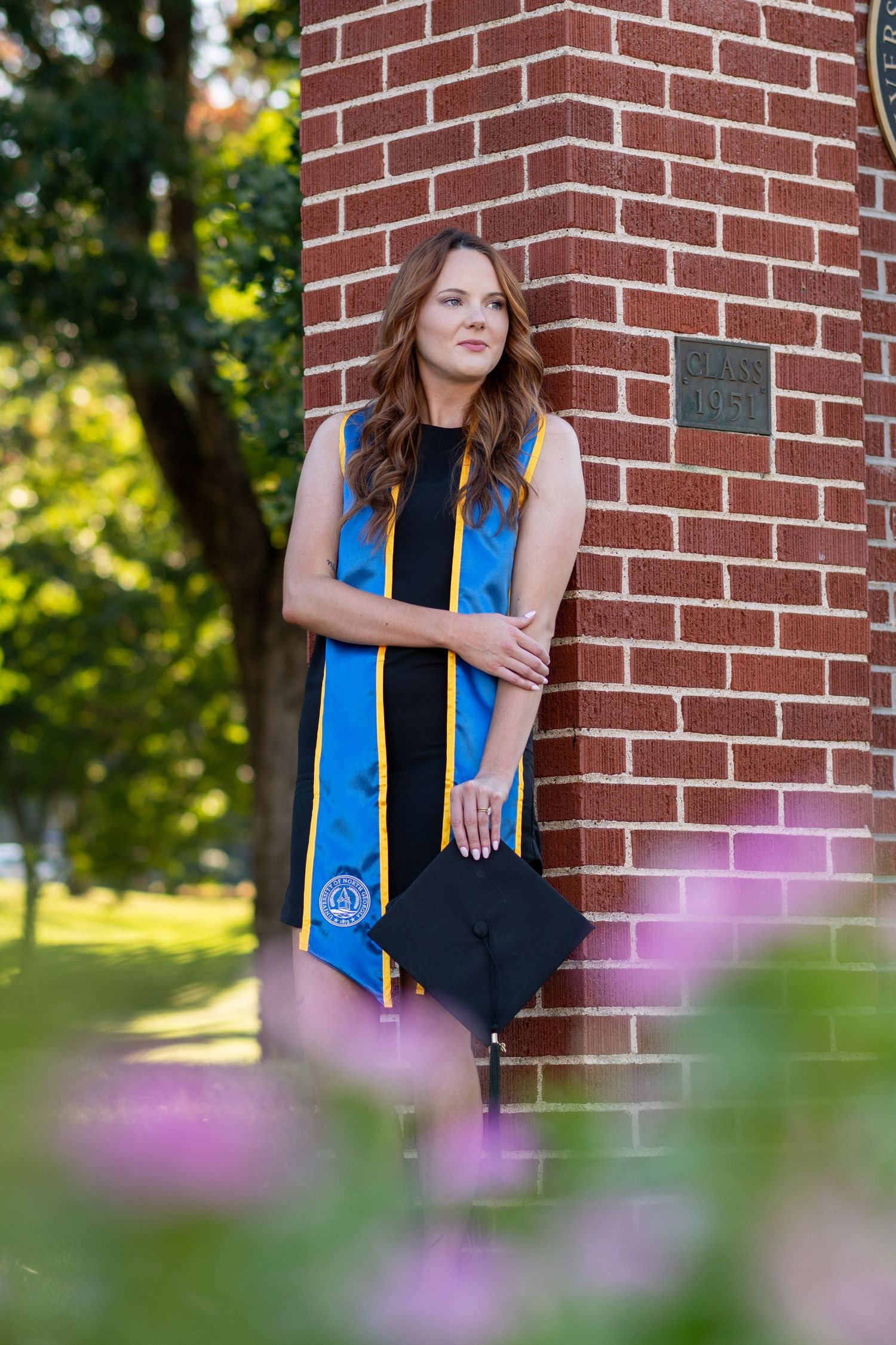 A young woman in graduation regalia with blue stole poses against a brick column outdoors.