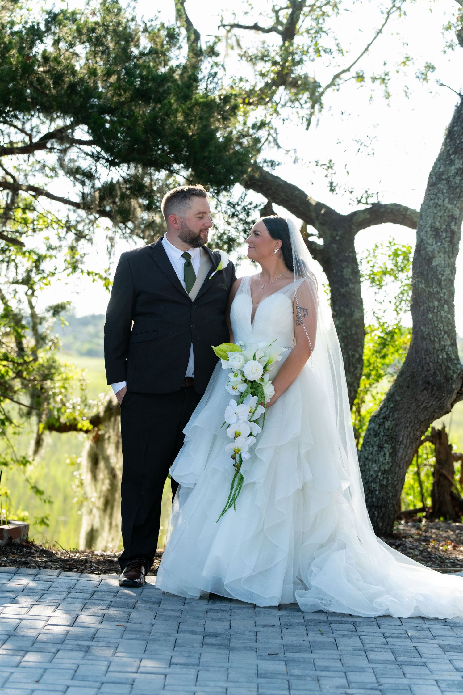 Bride and groom share a romantic moment outdoors under a large oak tree, bride holding white calla lily bouquet.