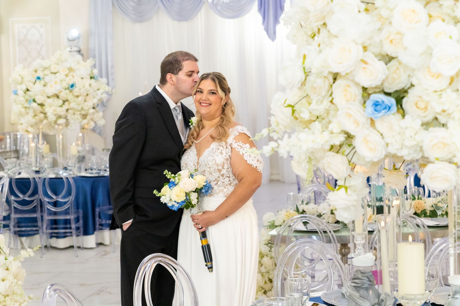 Bride and groom share a romantic kiss at elegant wedding reception with white floral arrangements and blue table settings.