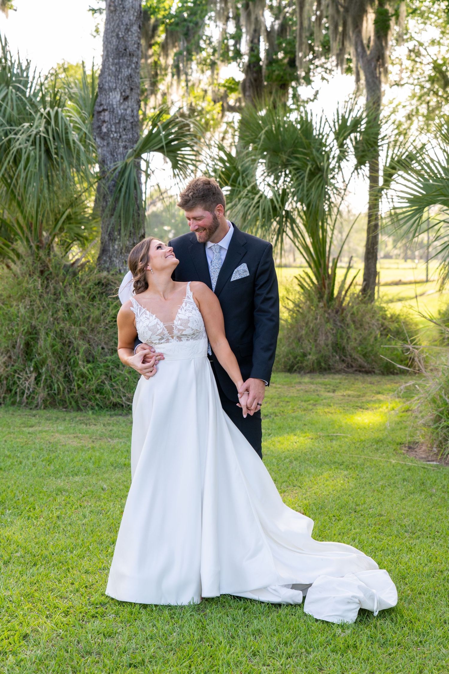 A bride and groom hold hands and share a romantic moment outdoors on green grass surrounded by palm trees.