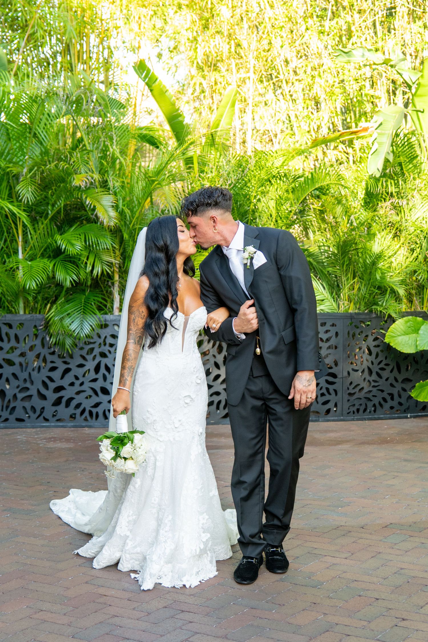 Bride and groom share a romantic kiss outdoors, surrounded by lush tropical greenery, bride holding white bouquet.