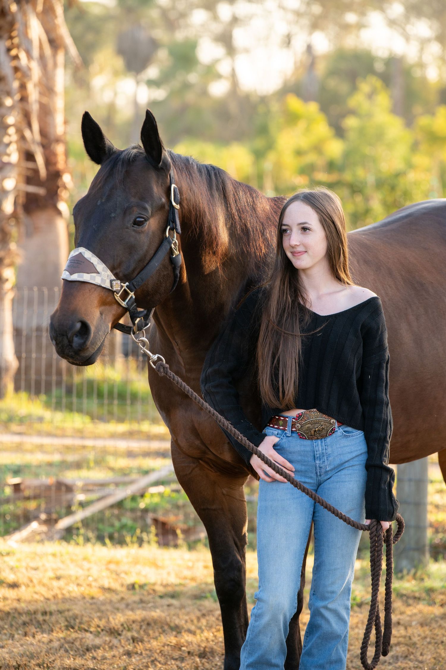 Young woman with long hair in jeans and black top stands beside a dark bay horse on a sunny farm.