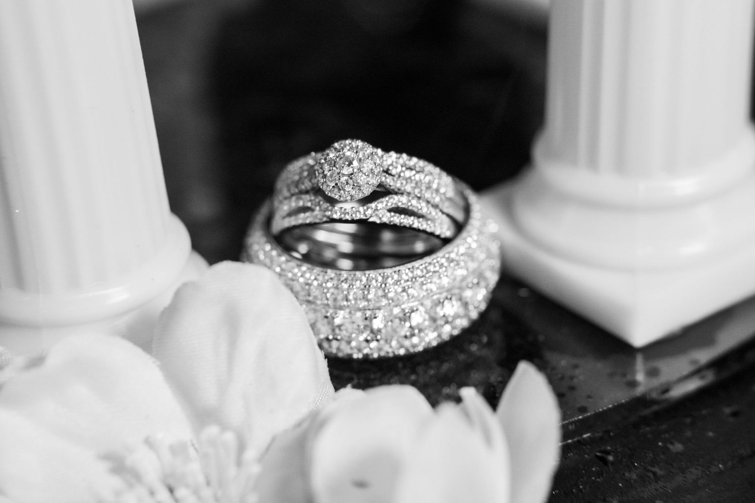 Black and white photo of sparkling diamond wedding ring set displayed with white flowers and decorative columns.