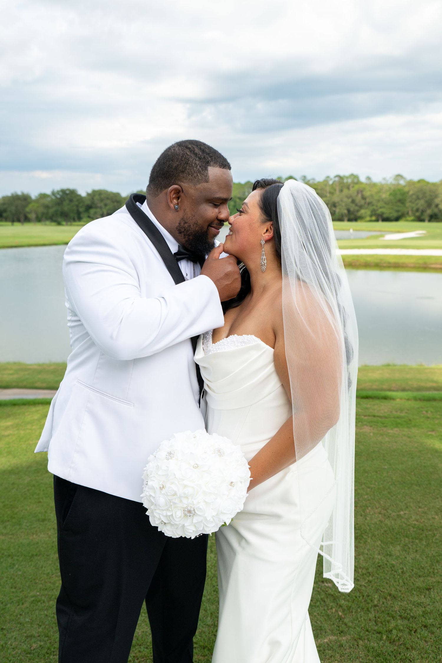 A bride and groom share a romantic moment on a golf course, with the bride holding a white bouquet and wearing a veil.