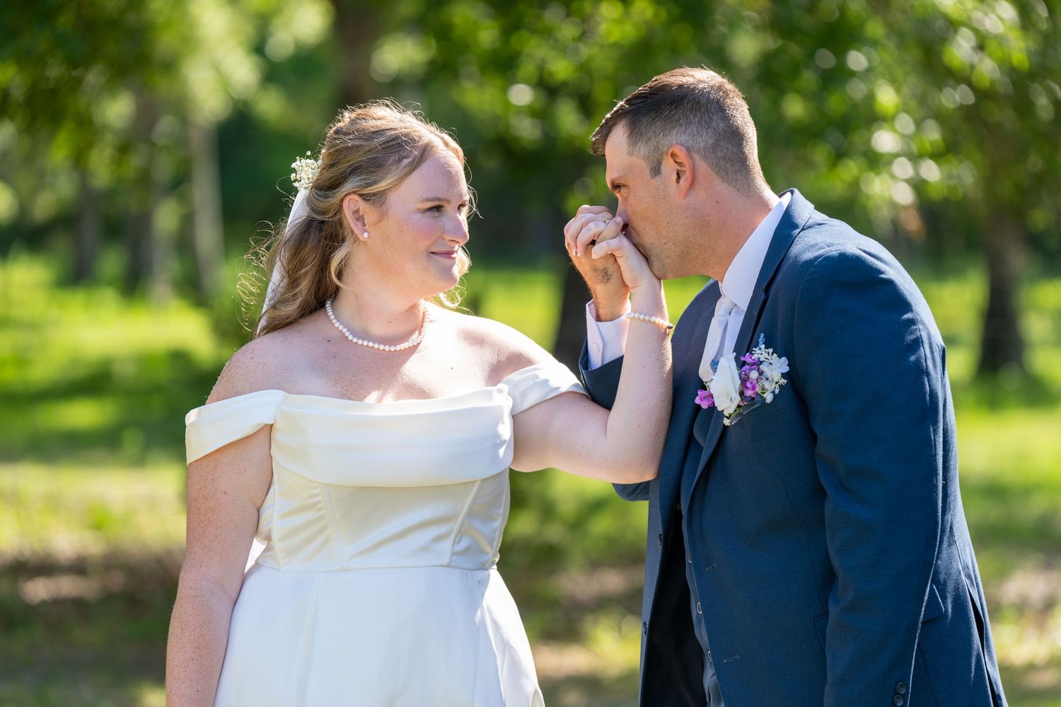 Groom in navy suit kisses bride's hand outdoors at wedding ceremony with lush green trees in background.