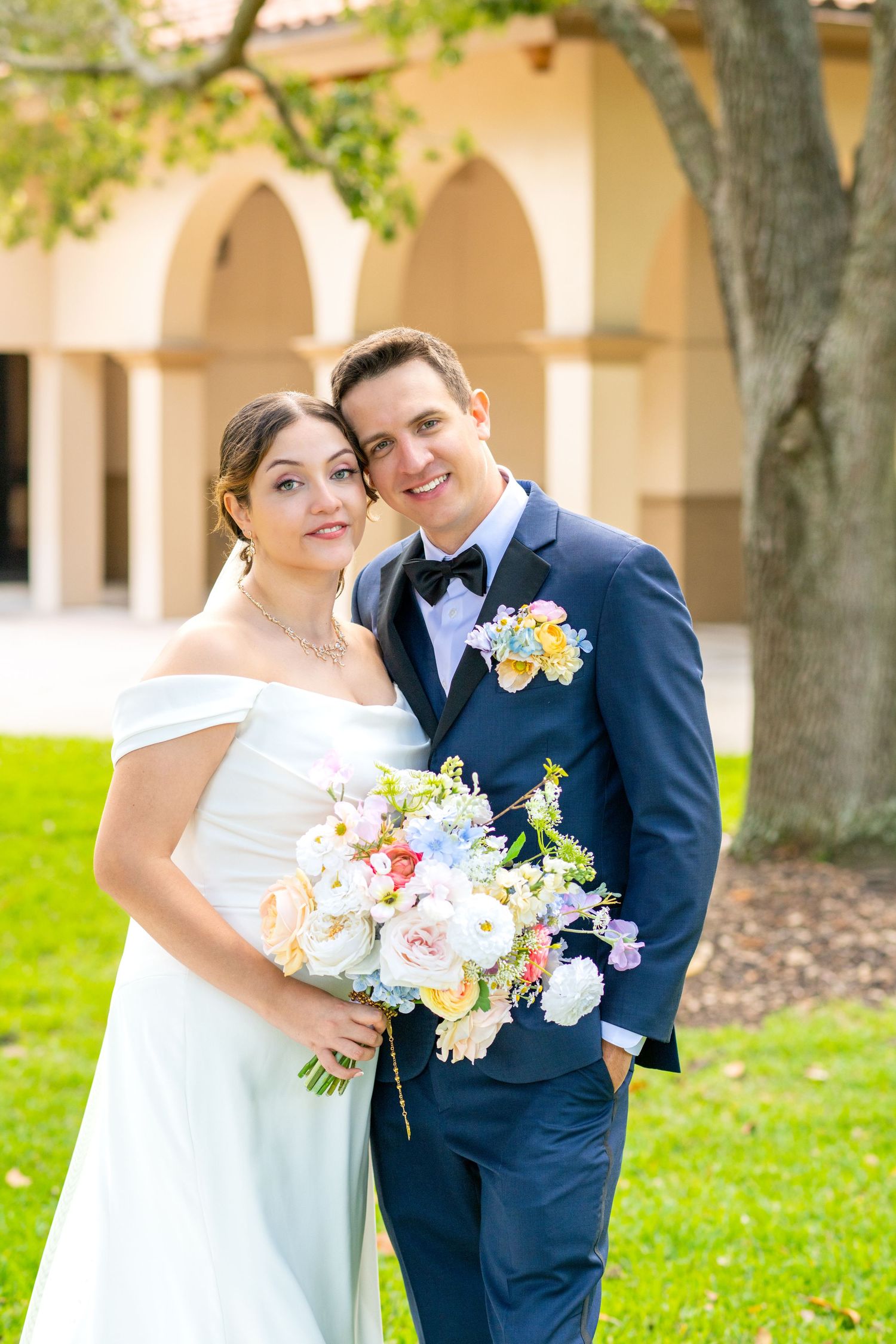Smiling bride and groom pose outdoors with a colorful spring bouquet near elegant arched architecture.