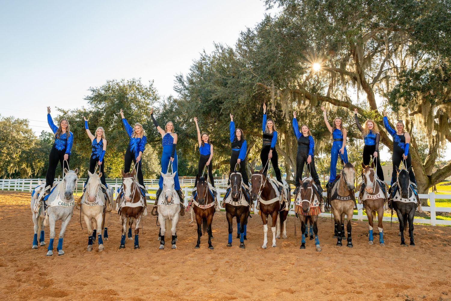 Group of equestrian riders in blue uniforms pose on horseback with arms raised at a sunny outdoor arena.