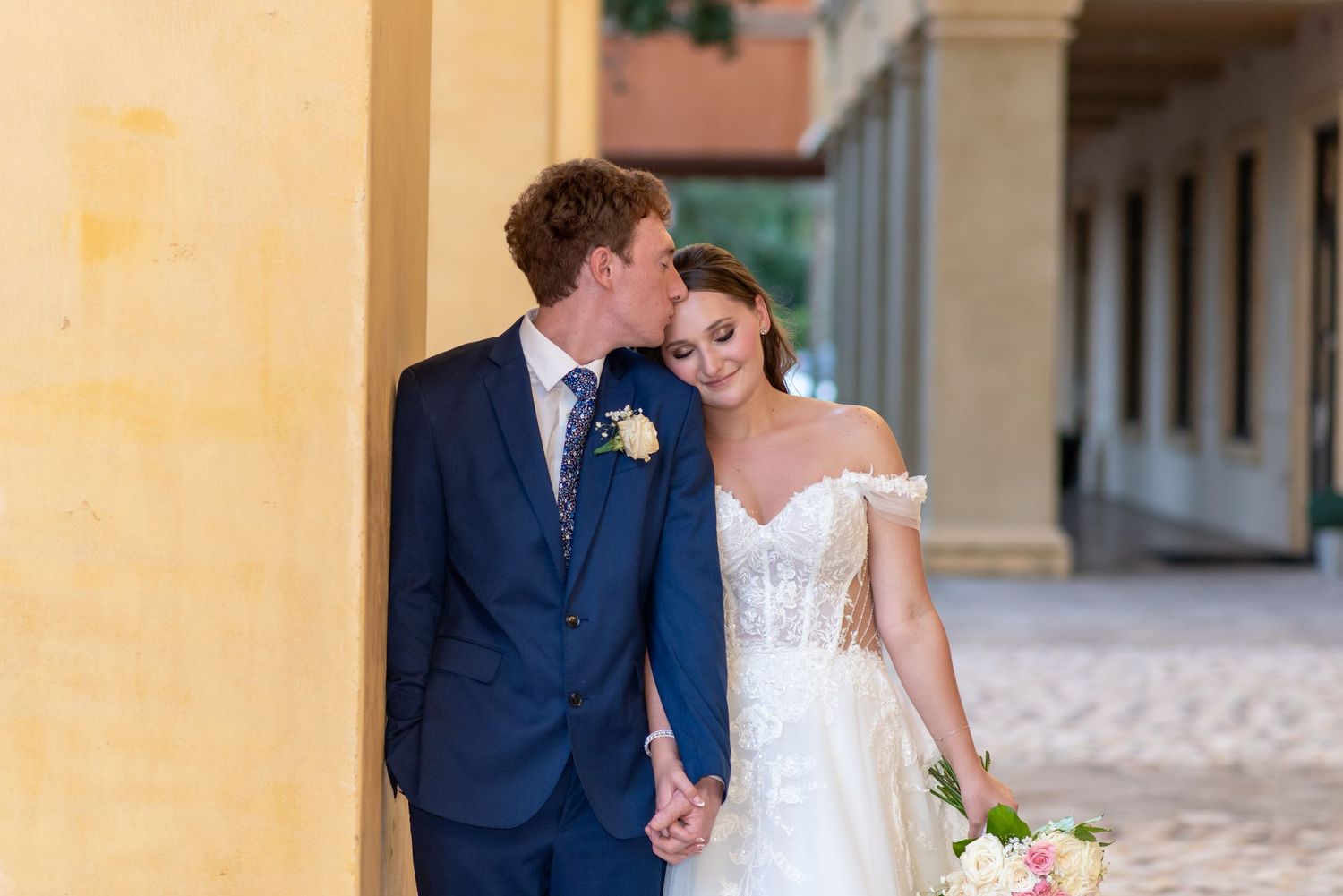 Bride and groom holding hands and sharing a romantic moment against a yellow column in an elegant outdoor setting.