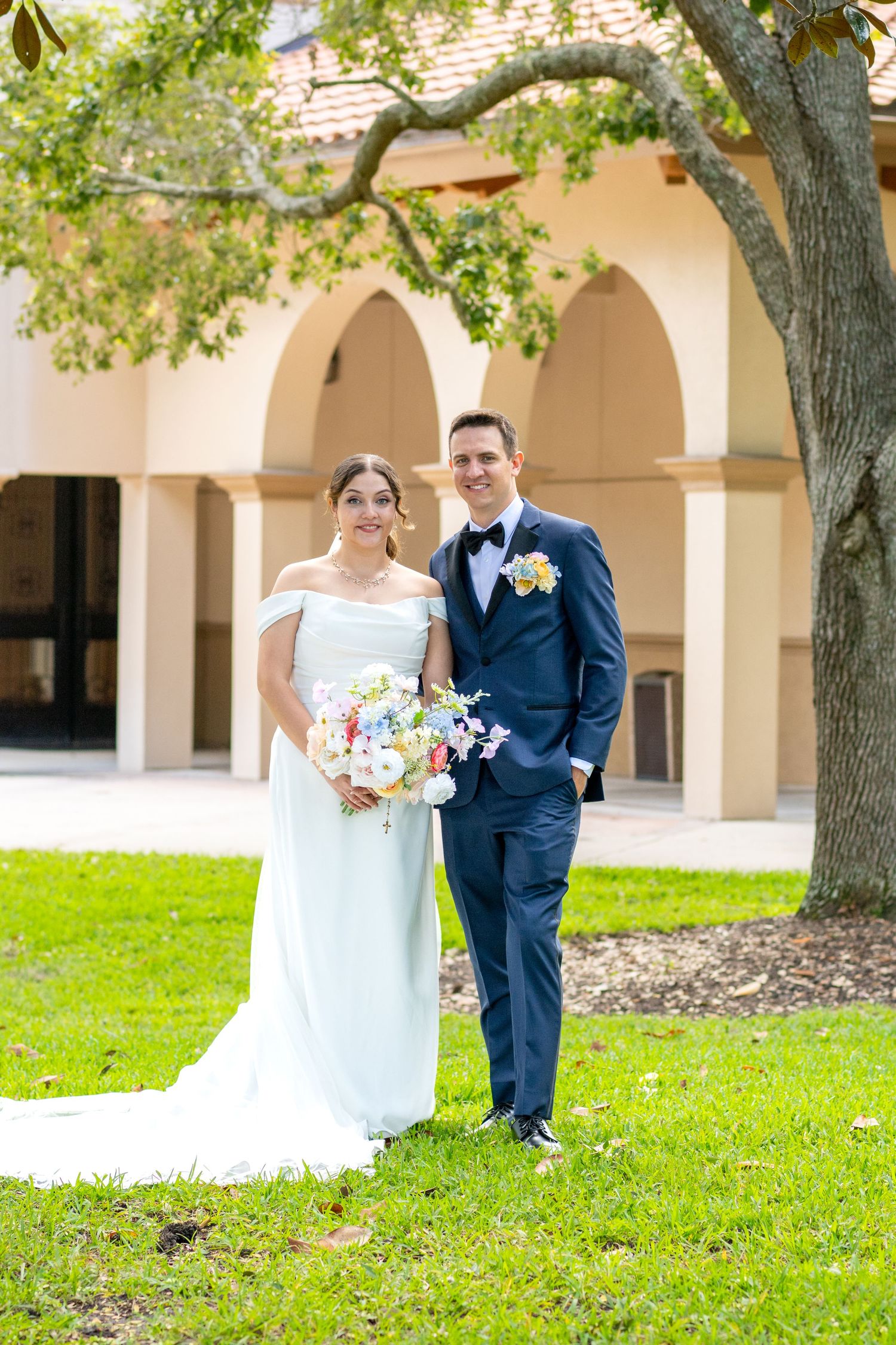 Bride in white off-shoulder gown and groom in navy suit pose together on green lawn before arched building.
