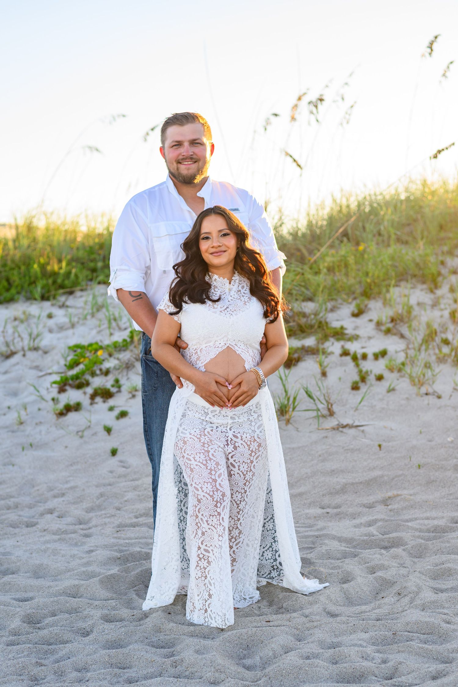 Expectant couple in white outfits pose on a sandy beach with sea oats at sunset, the woman cradling her baby bump in a lace gown.