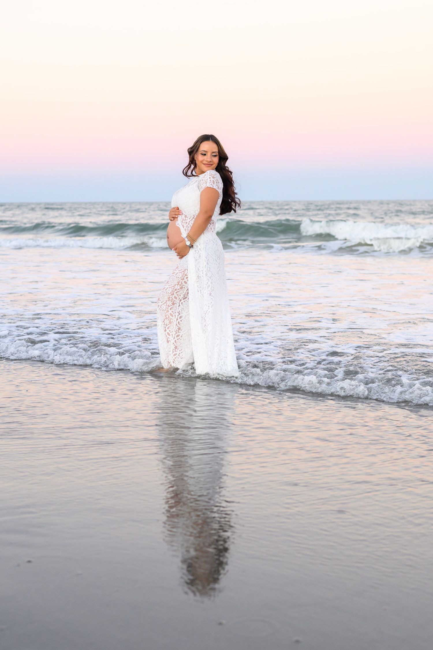 Pregnant woman in a flowing white lace dress cradles her baby bump while standing in shallow ocean waves at sunset, with a pink sky.