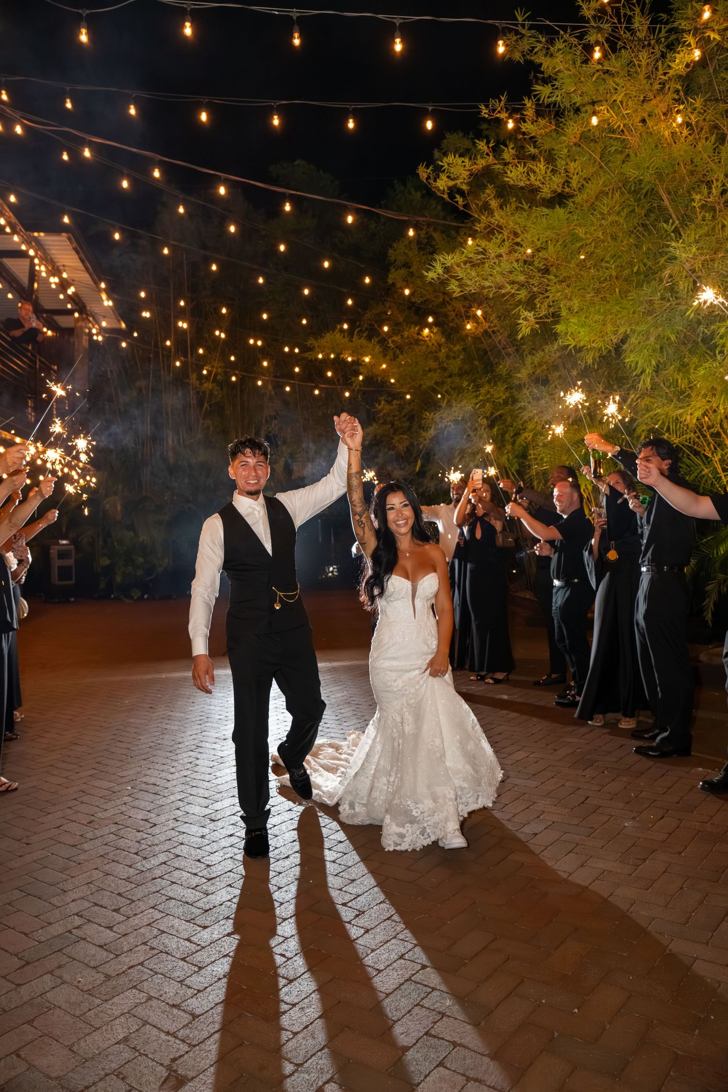 Bride and groom exit wedding reception under string lights with sparklers lining the path at night.