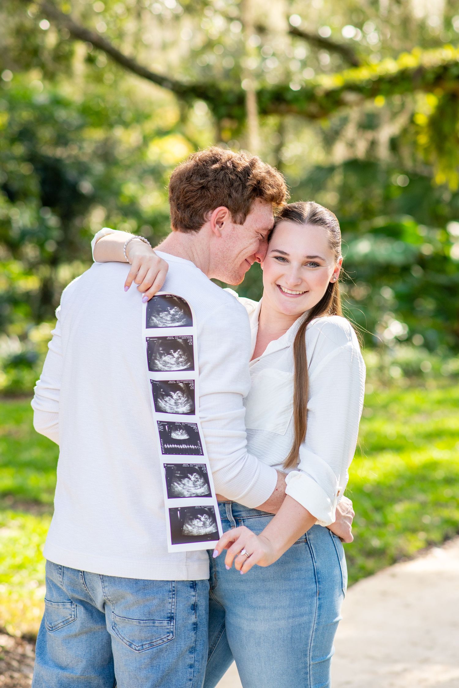 Happy couple embracing outdoors holding ultrasound photos, announcing pregnancy in a sunny green park setting.