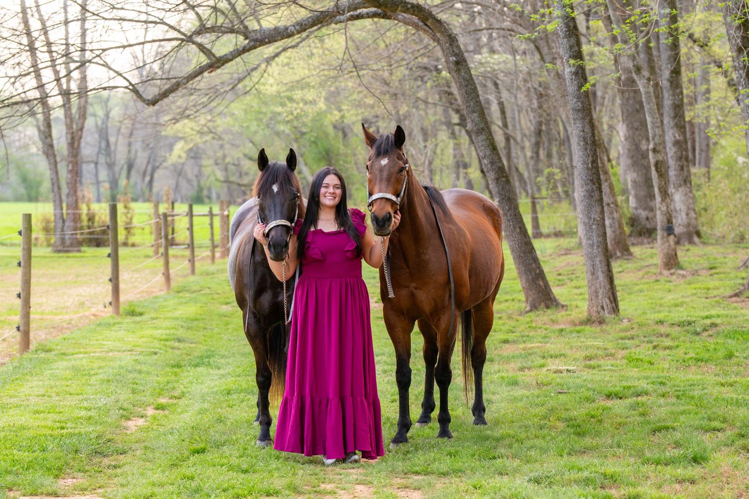Woman in magenta gown poses with two horses on green pasture surrounded by trees.