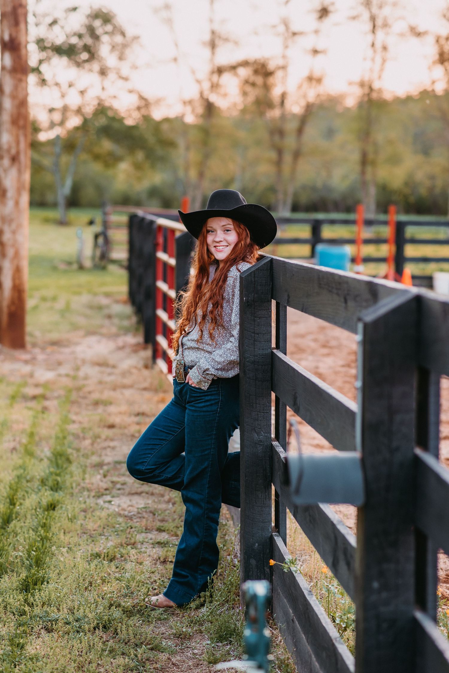 Young woman in black cowboy hat and jeans leaning on wooden fence at golden hour on a ranch.