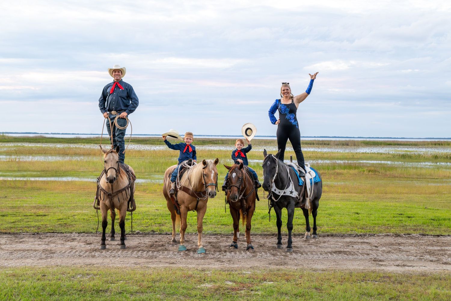 Four performers in western attire stand and balance on horseback in an open field near water under a cloudy sky.