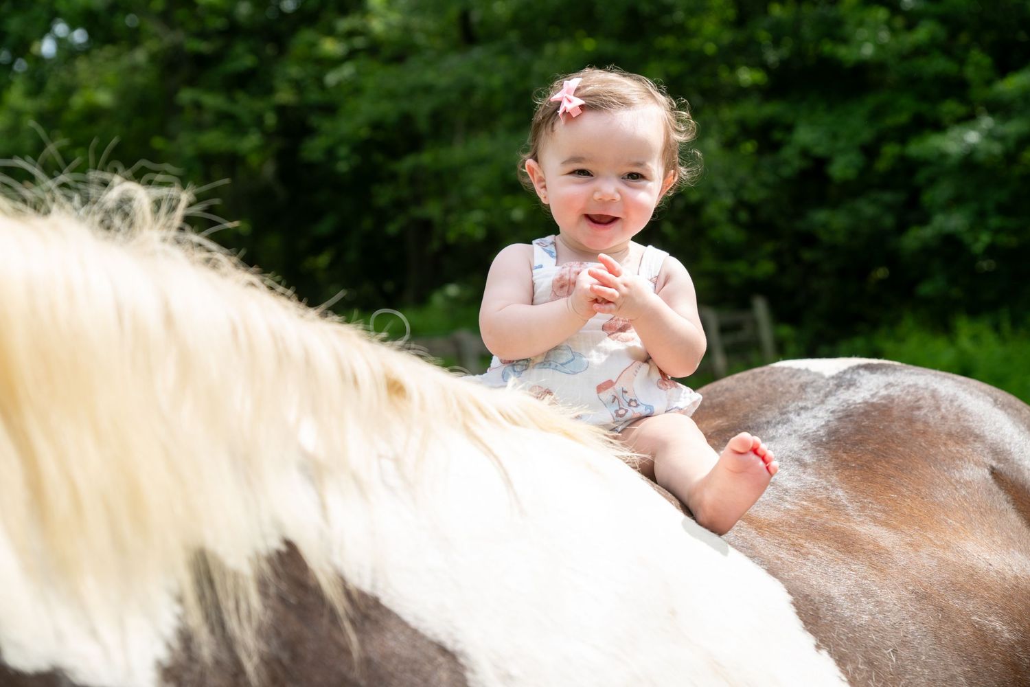 Happy baby girl sitting on a horse's back outdoors, smiling joyfully in a floral romper on a sunny day.