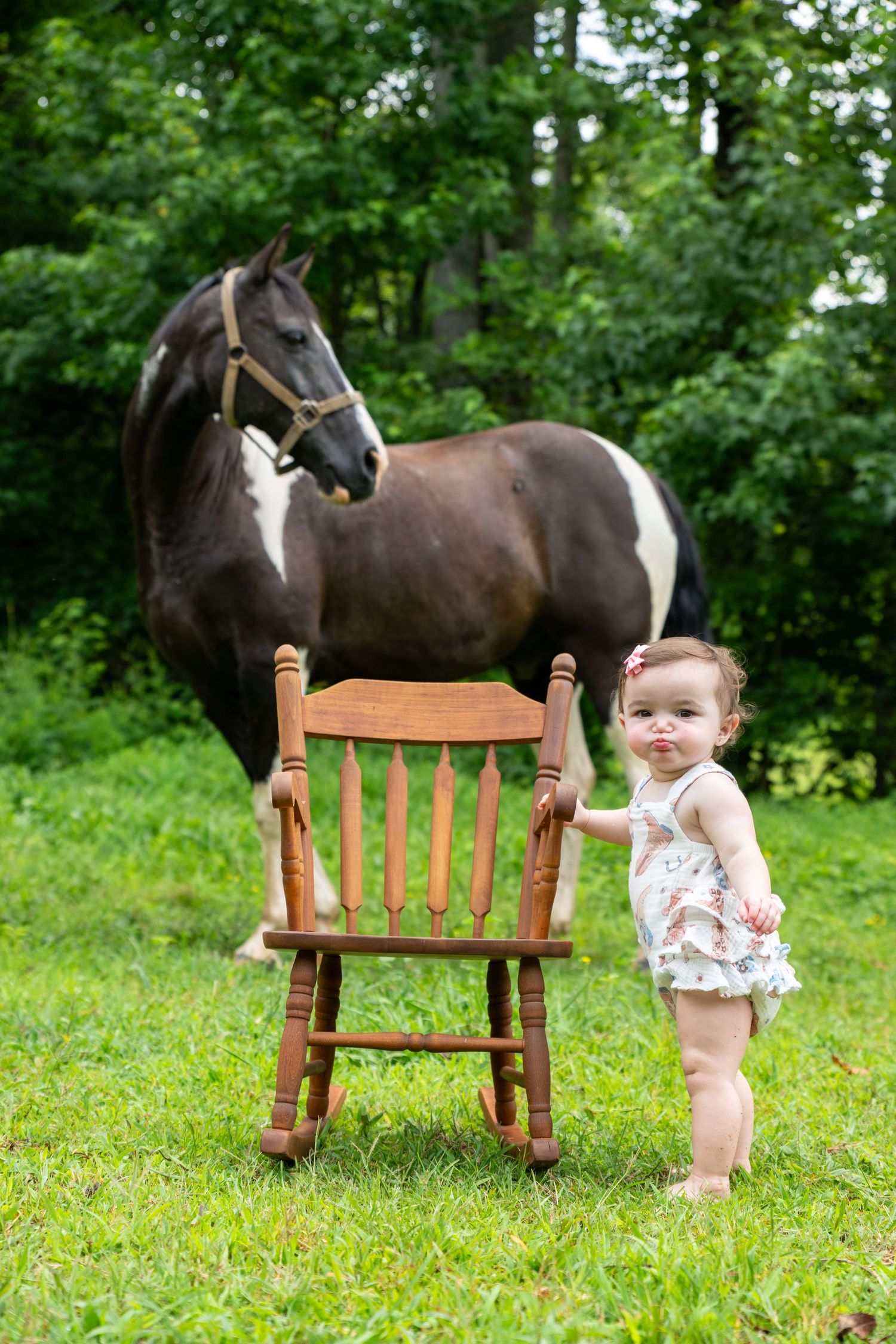 Adorable baby girl standing by a wooden rocking chair with a paint horse in a lush green field.