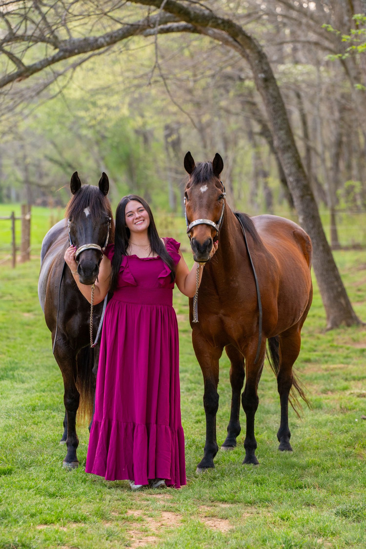 Smiling young woman in magenta gown standing between two bay horses on green grass with trees in background.