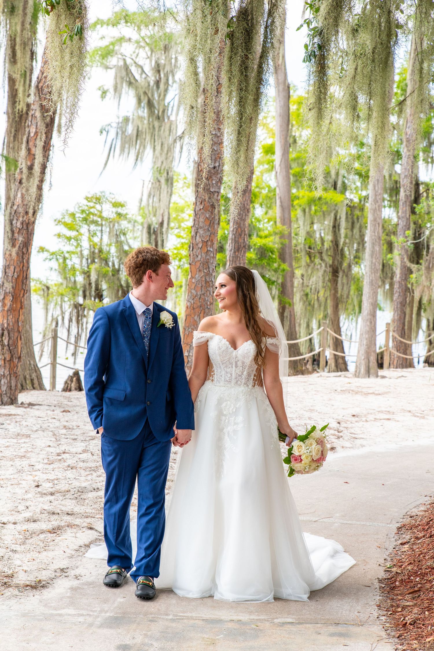 Bride and groom walk hand-in-hand under Spanish moss-draped trees, she in a white gown, he in a navy suit.