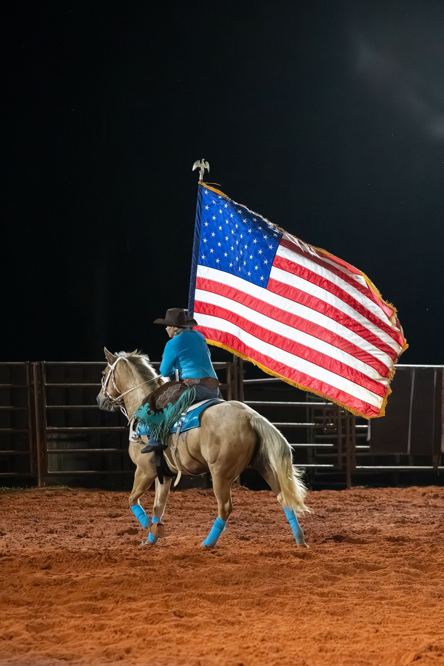 Rider in turquoise shirt on palomino horse carrying large American flag in dark rodeo arena.