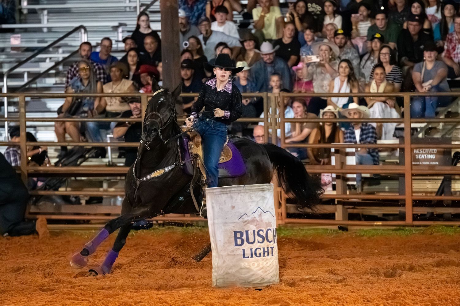 Rider on a black horse performs barrel racing near a Busch Light barrel at a packed indoor rodeo arena.