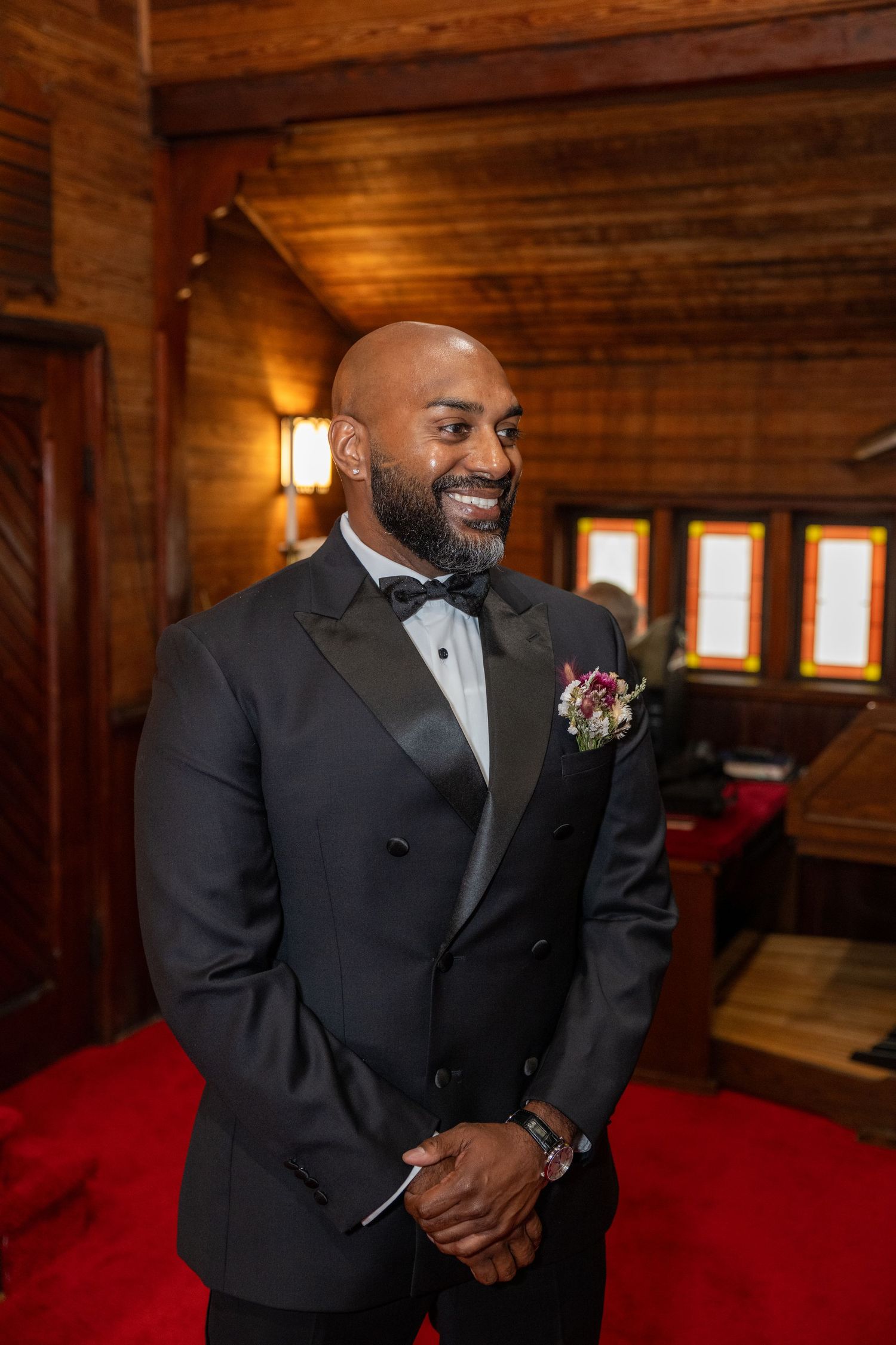 Smiling bald groom in black tuxedo with boutonniere standing in a warmly lit wooden chapel interior.