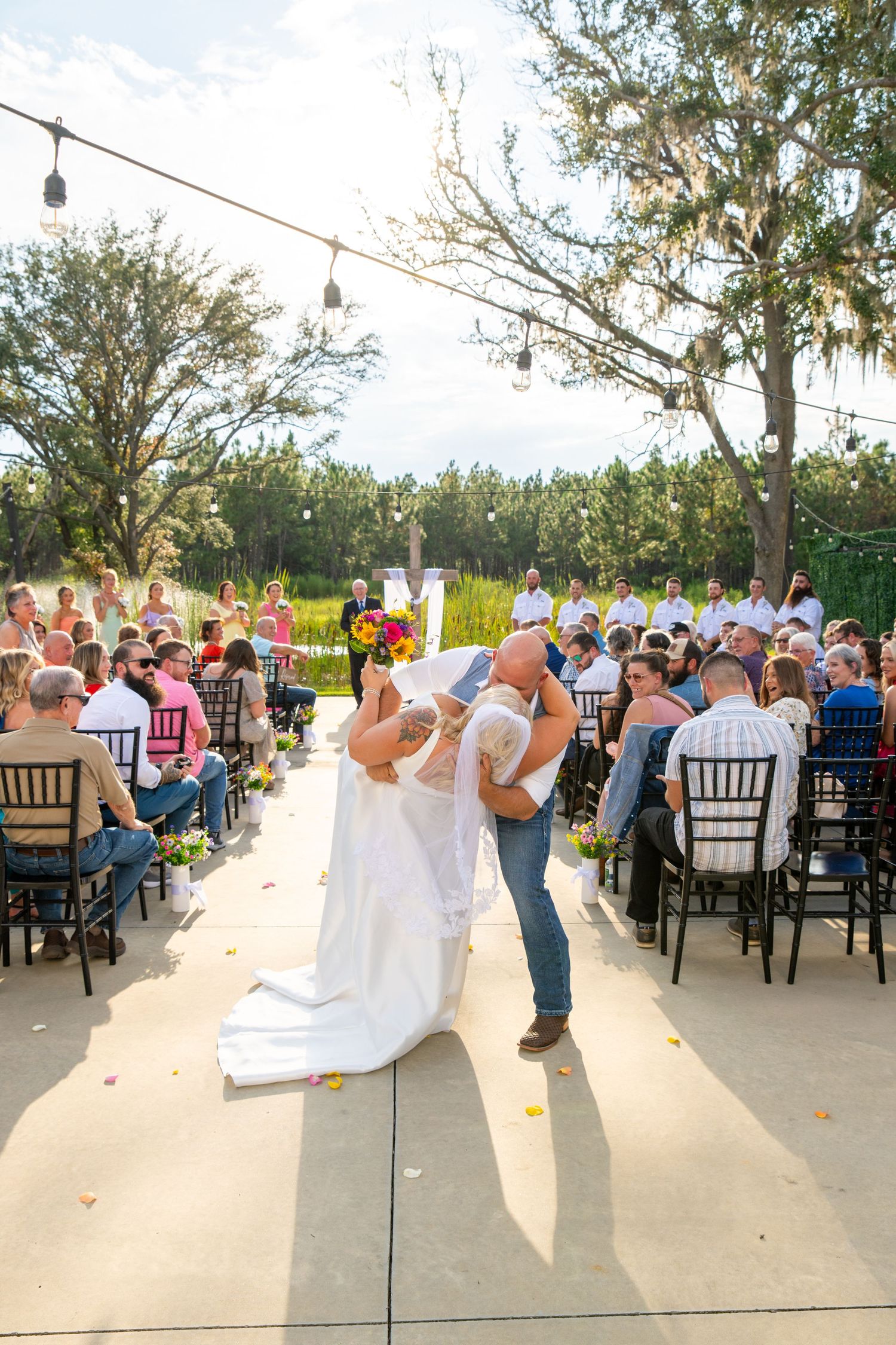 Groom dipping bride at outdoor wedding ceremony aisle with seated guests.