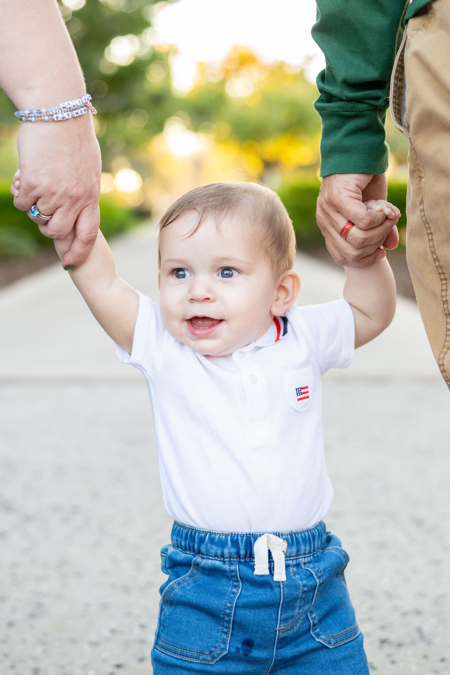 Adorable blue-eyed baby boy standing and holding parents hands on outdoor pathway, smiling brightly.