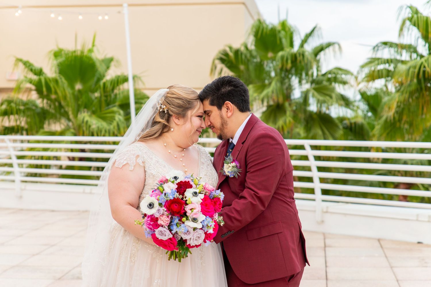 Bride and groom touching foreheads tenderly, she holds vibrant pink bouquet, he wears burgundy suit, tropical palm trees background.