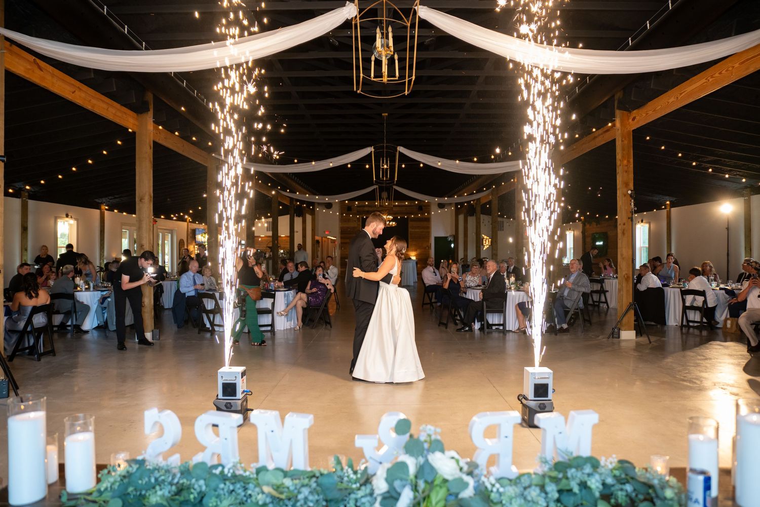 Couple shares first dance surrounded by sparkling cold sparks at elegant barn wedding reception with string lights.