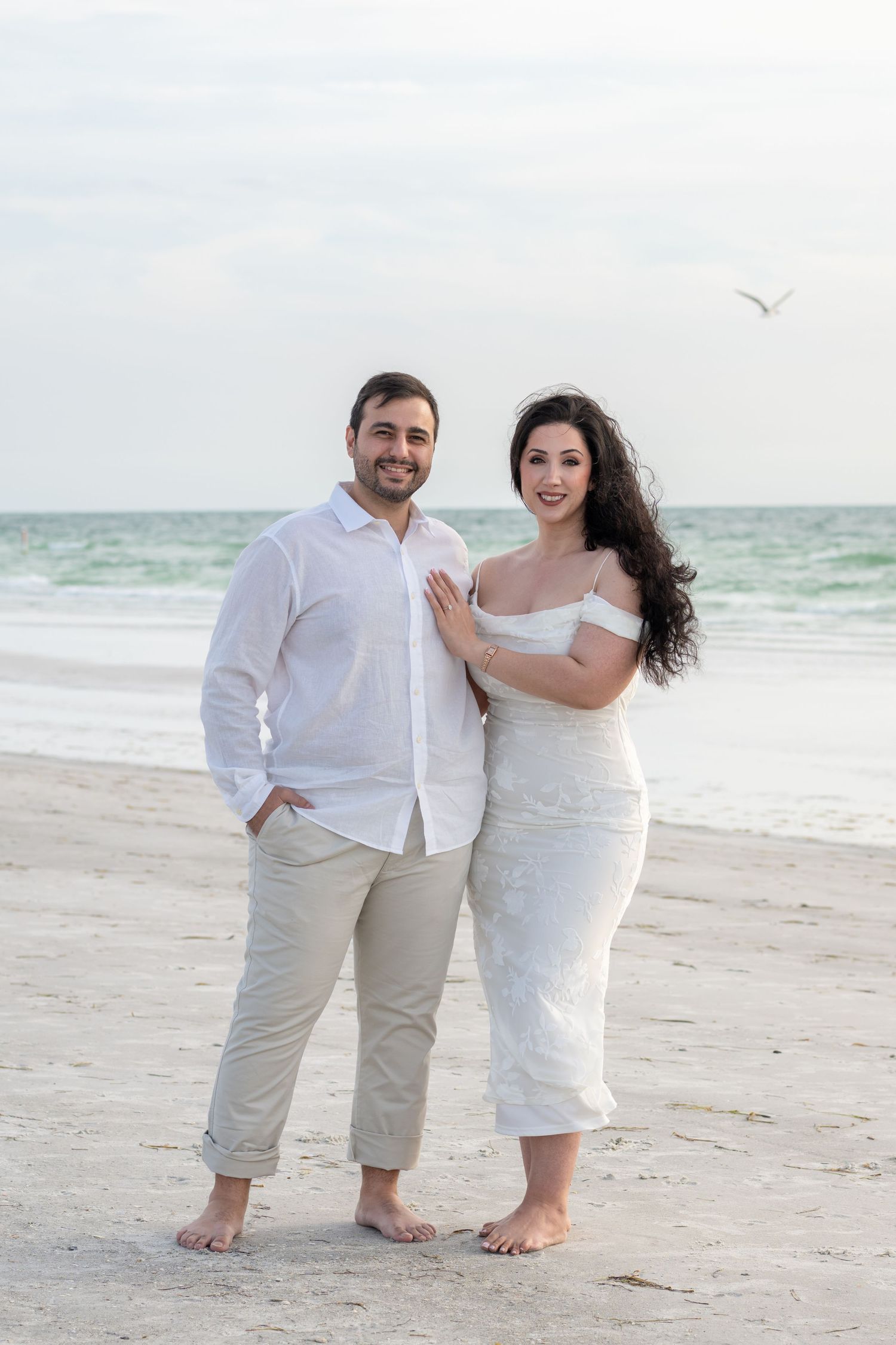 Couple in white outfits posing barefoot on a sandy beach with ocean waves in the background.