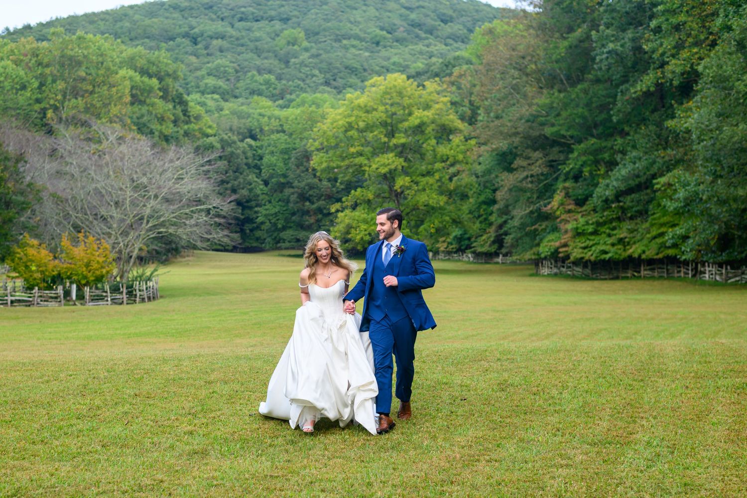 Bride and groom walking hand in hand across a lush green meadow with rolling forested hills in the background.