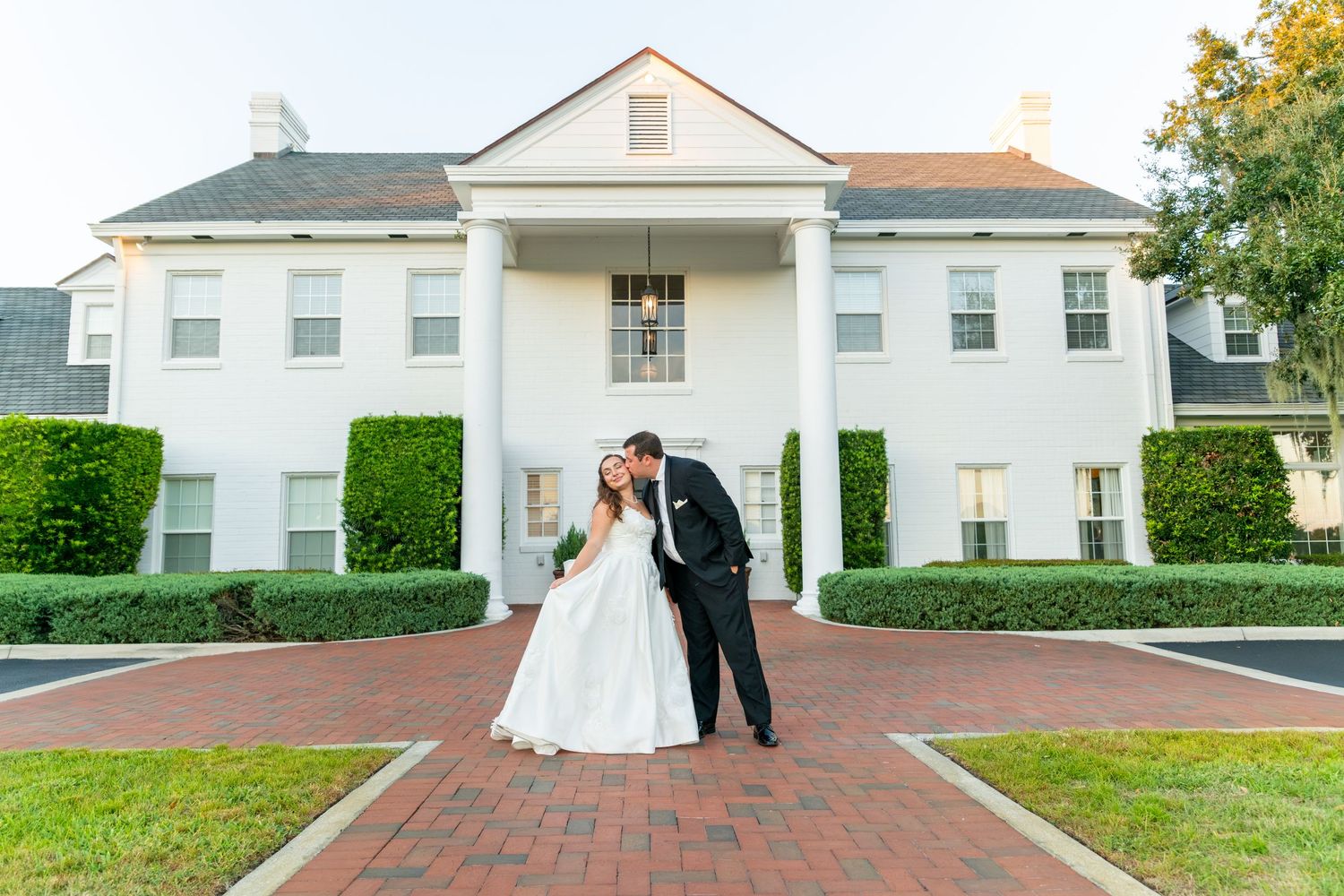 Bride and groom share a romantic kiss in front of a grand white colonial wedding venue with brick pathway.