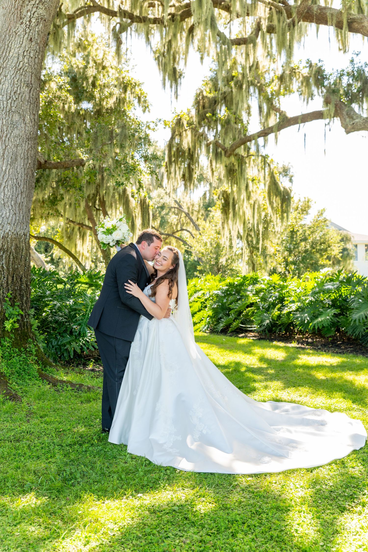Bride and groom share a romantic kiss under a moss-draped oak tree on lush green grass at an outdoor wedding.