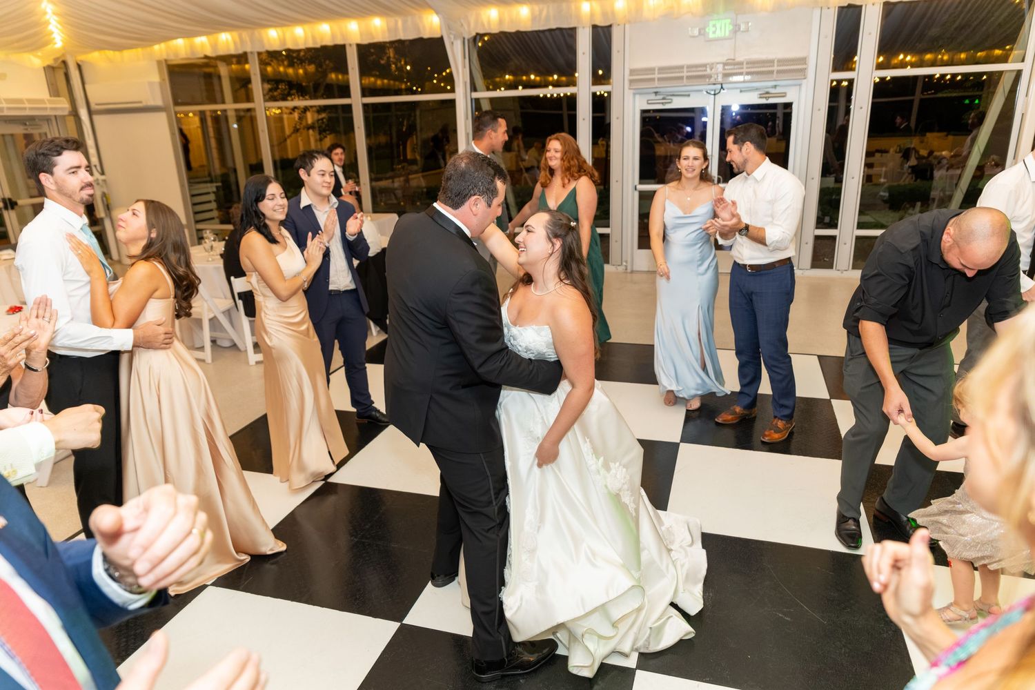 Bride and groom share first dance on black and white checkered floor surrounded by wedding guests at elegant reception.
