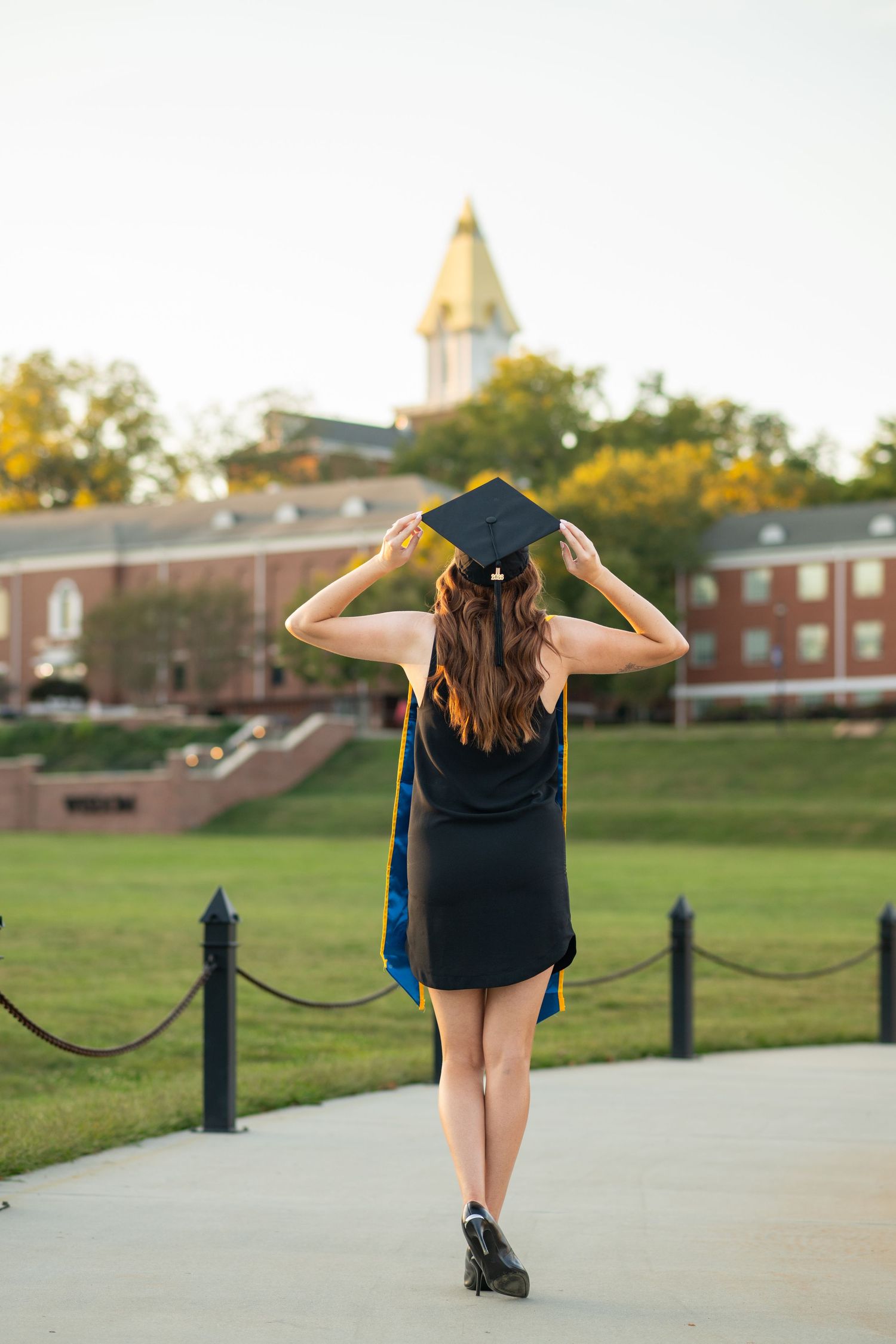 A woman in a black dress and graduation cap walks along a campus pathway toward a brick building with a gold dome.